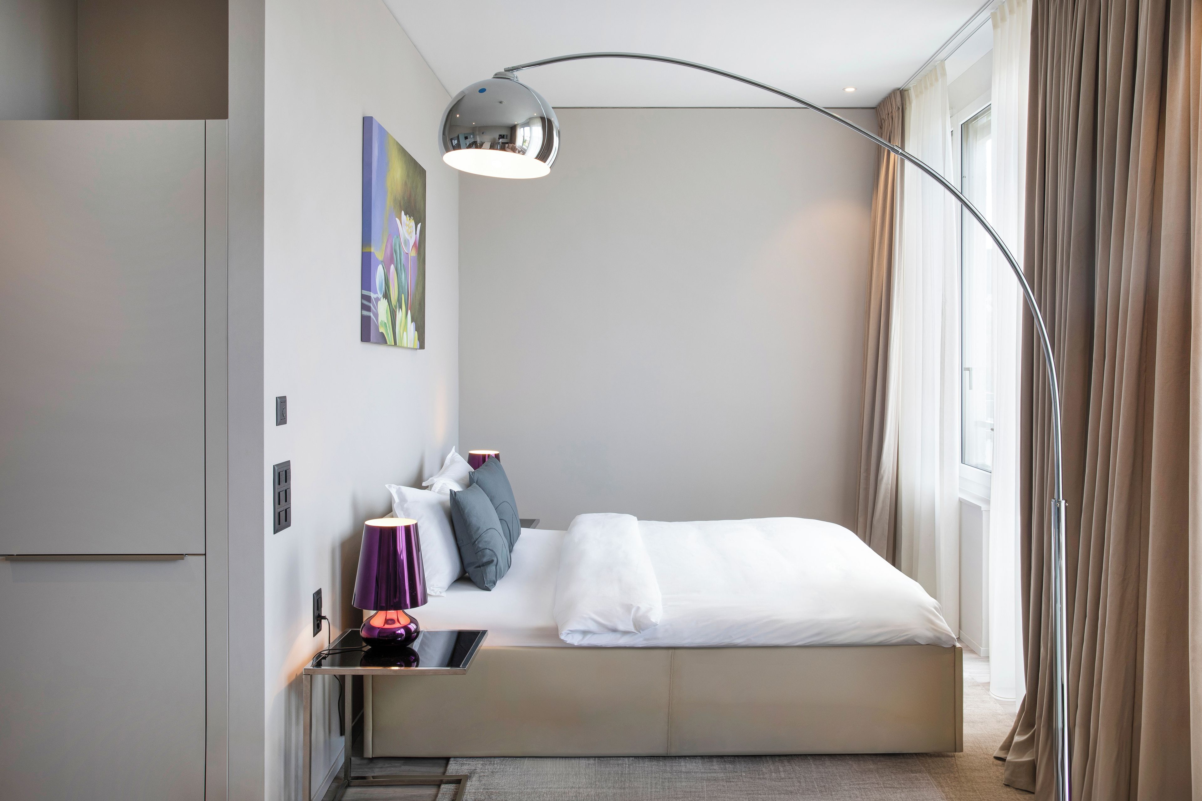 Une chambre d’hôtel moderne équipée d’un lit, d’une table de chevet, d’une lampe et d’une grande fenêtre avec des rideaux.