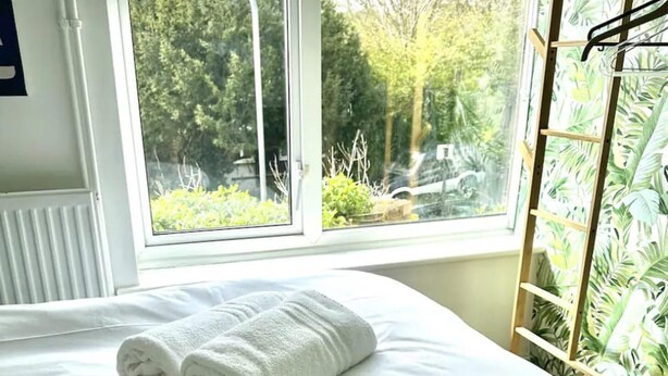 A neatly made bed with a rolled white towel, two small bottles, and a ladder against a window with a view of greenery outside. - Deluxe Beach House Brighton (Brighton)