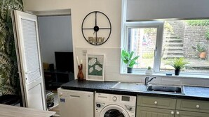 A compact kitchen with a washing machine, a dining area with stools, and a window with a view of a brick wall and plants outside. - Deluxe Beach House Brighton (Brighton)