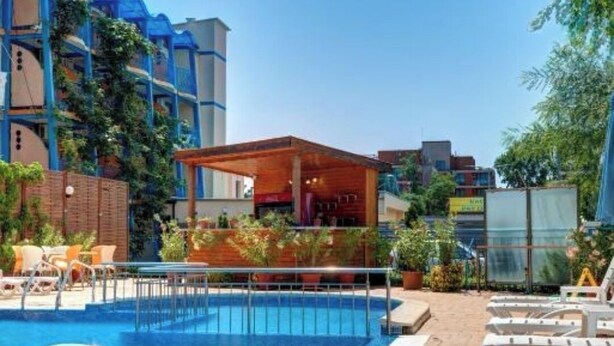 A swimming pool with a wooden deck and lounge chairs, surrounded by buildings with blue and white facades. - Hotel Paloma (Sunny Beach)