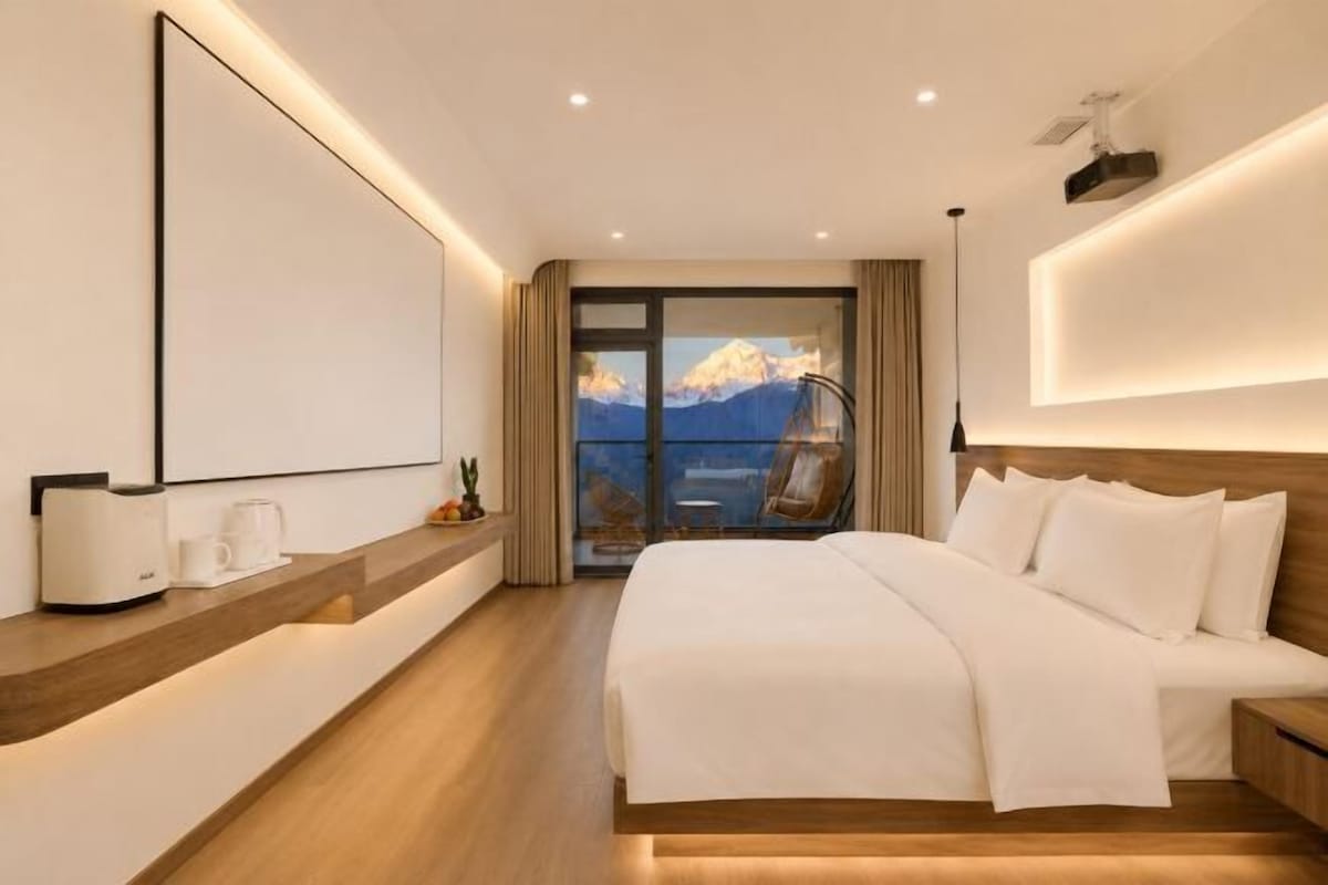 A hotel room with a large bed, a bedside table, a mirror, and a view of mountains through a glass door.