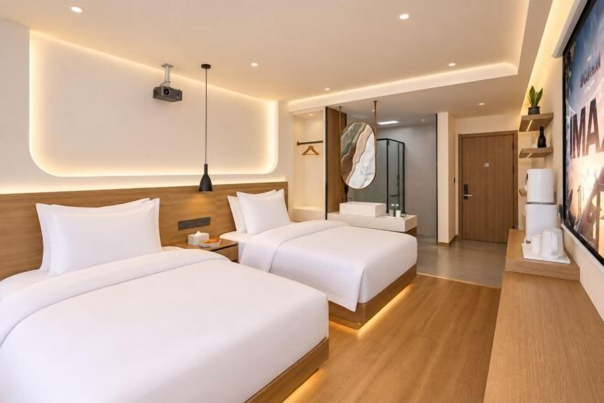 A hotel room with two beds, a bedside table, a lamp, a mirror, a bathroom door, and a poster on the wall.