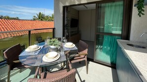 Un balcon avec une table en verre dressée pour le dîner, des chaises en osier, une petite plante en pot et une vue sur un toit recouvert de tuiles rouges.