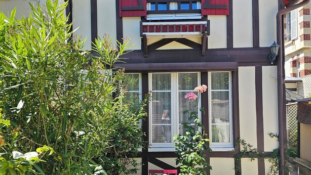 Une maison traditionnelle à colombages, avec une façade rouge et blanche, un petit balcon et un jardin agrémenté d’une table et de chaises.