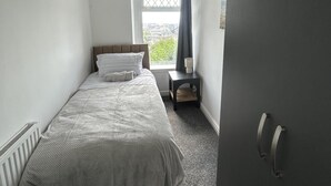 Piccola camera con un letto singolo, una finestra, un comodino con una lampada e un termosifone.