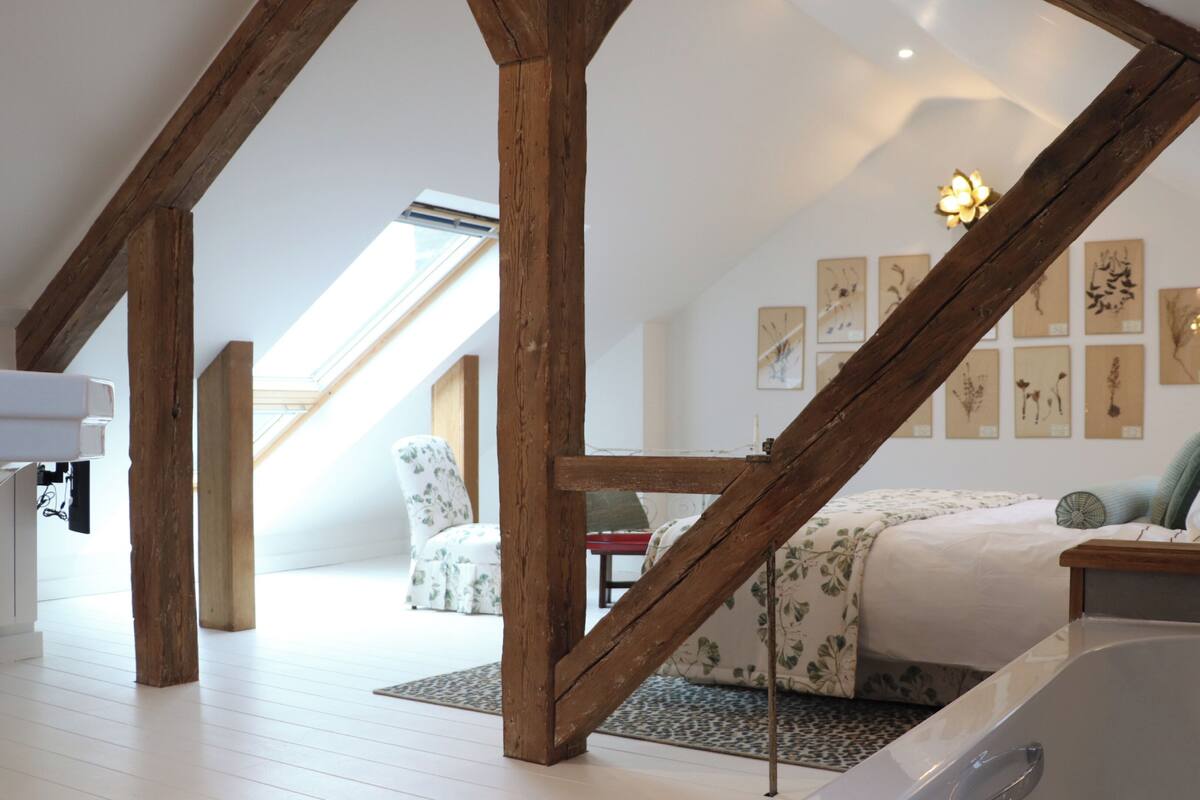 A spacious attic bedroom with exposed wooden beams, a skylight, a bed with a floral bedspread, a small table, and a wall with framed botanical prints.