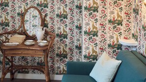 Una habitación con papel tapiz estampado, una silla de mimbre, una mesita con tazas de té y un sofá color verde azulado con un cojín.