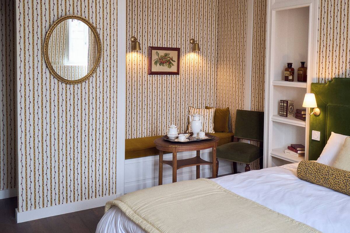 A hotel room with a bed, a small table with tea set, a chair, a shelf with books, and a wall with vertical lines.
