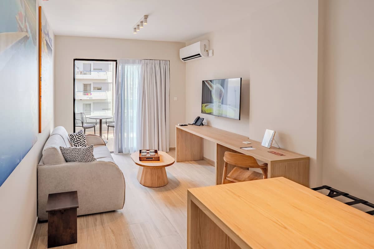 A modern, well-lit living room with a sofa, a wooden coffee table, a dining table, and a TV mounted on the wall.