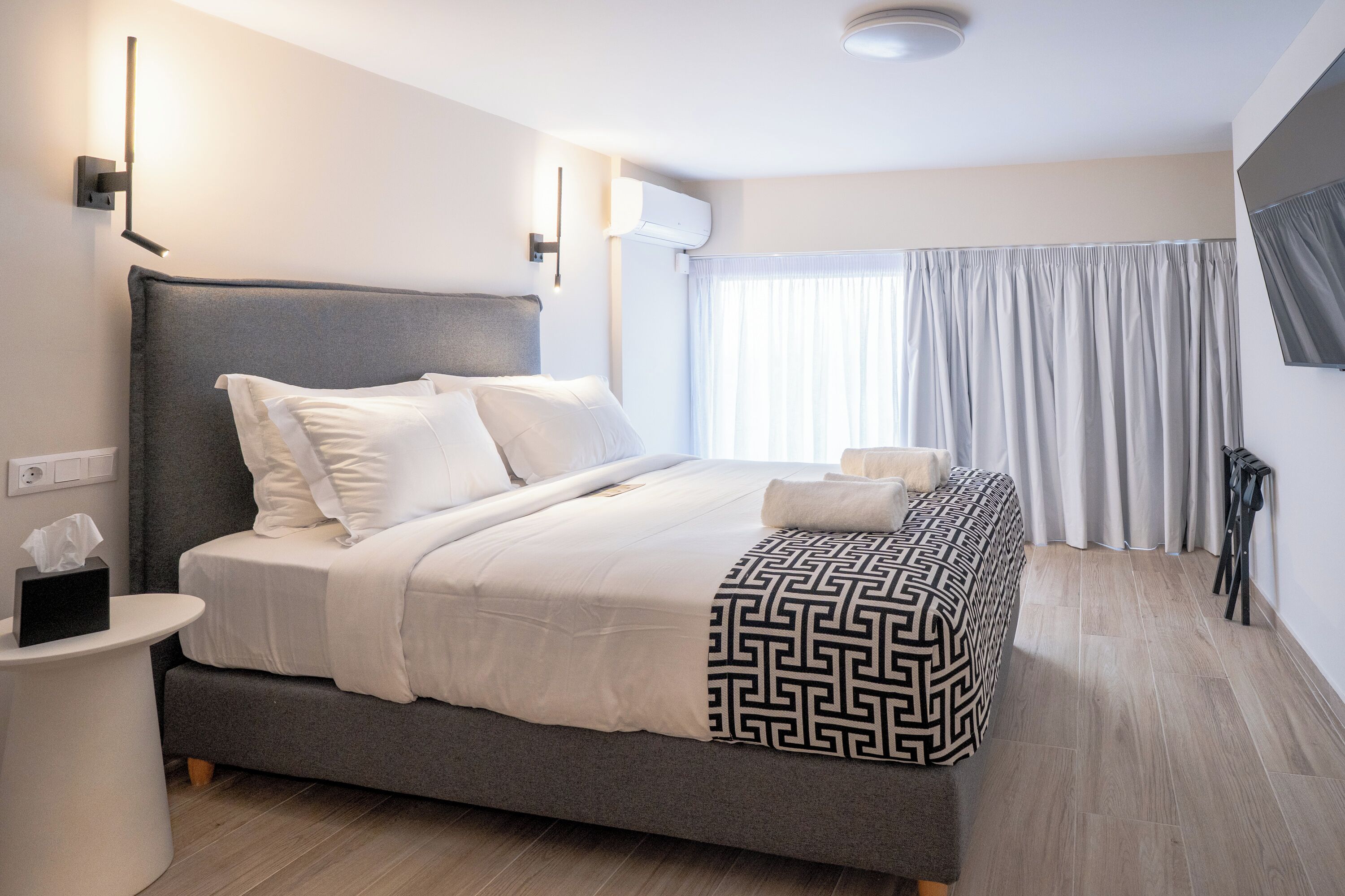 A modern hotel room with a large bed, a bedside table, a wall-mounted air conditioner, and a window with curtains.