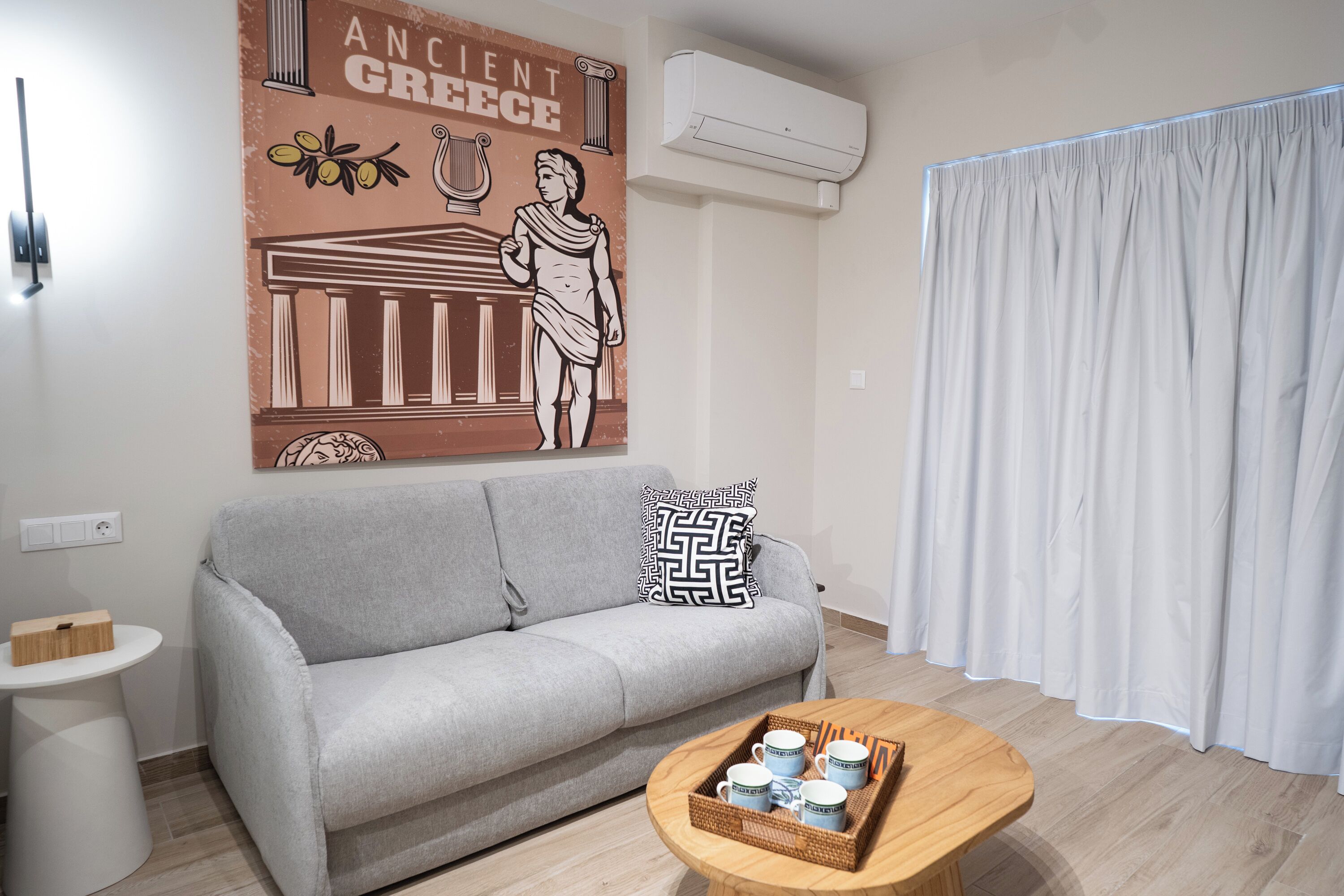A modern living room with a grey sofa, a coffee table with cups, a wall-mounted air conditioner, and a large decorative poster.