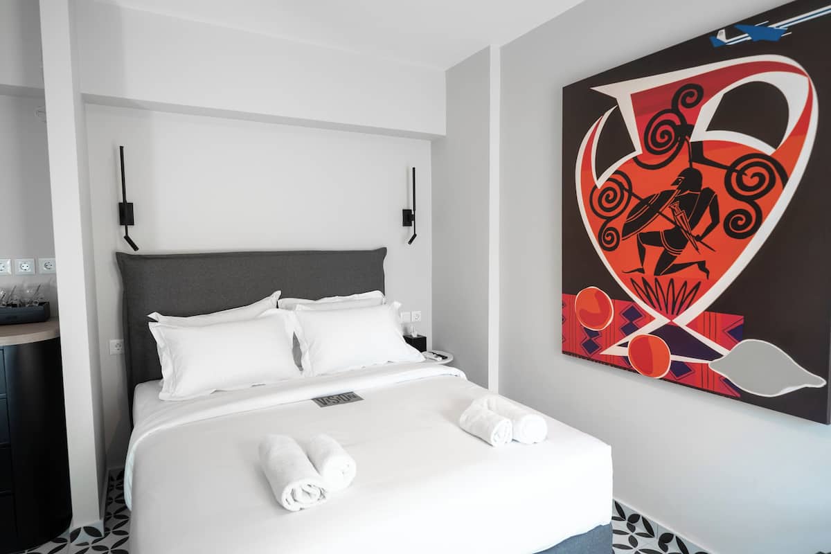A hotel room with a bed, white linens, a grey headboard, a wall-mounted artwork, and a bedside table with a phone and a lamp.