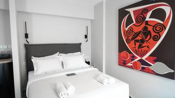 A hotel room with a bed, white linens, a grey headboard, a wall-mounted artwork, and a bedside table with a phone and a lamp.