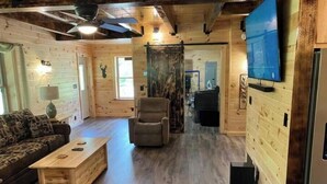 A cozy cabin interior with a wooden floor, a sofa, a chair, a small table, a television mounted on the wall, and a ceiling fan. - Creekside Cabin with Lake Access , Brand new build !!! (Remsen)