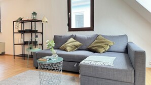 A modern living room with a grey sofa, a small round side table, a metal wire shelf, and a window with a view of a building. - Erholung pur am Rhein (Sankt Goar)