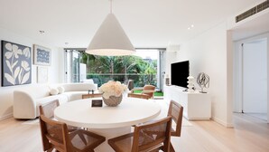 A modern living room with a round dining table, white sofas, and a large window overlooking a garden.