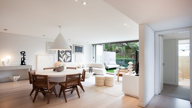A modern living room with a dining area, featuring a round table, wooden chairs, a white sofa, and a large window offering a view of the outdoors.