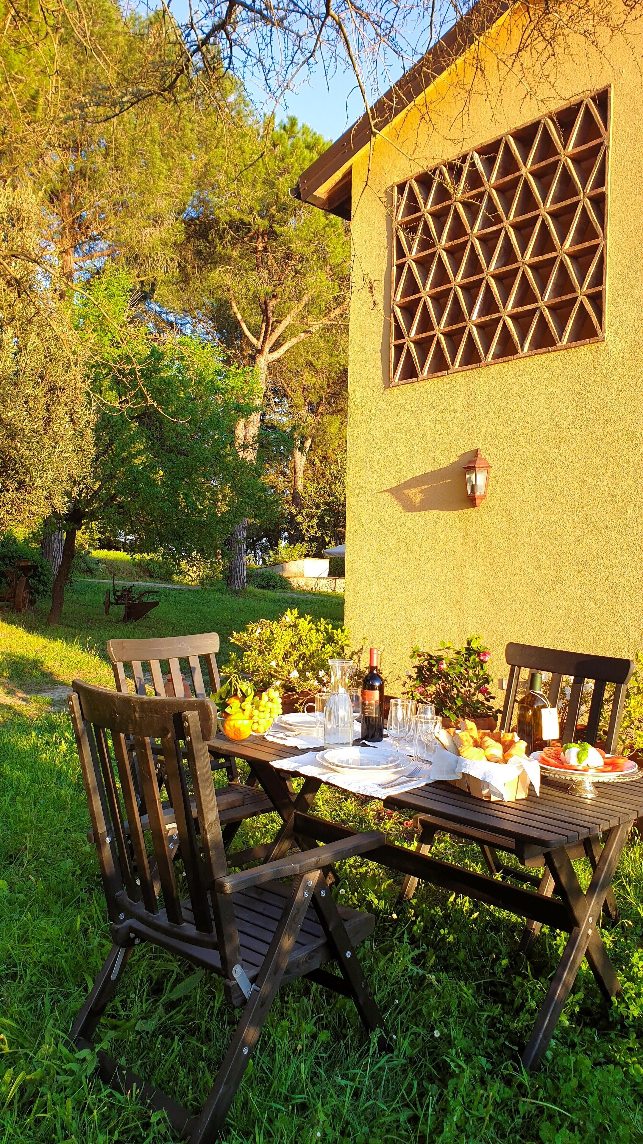 An outdoor dining area with a table set for two, surrounded by greenery and a building with a lattice window.