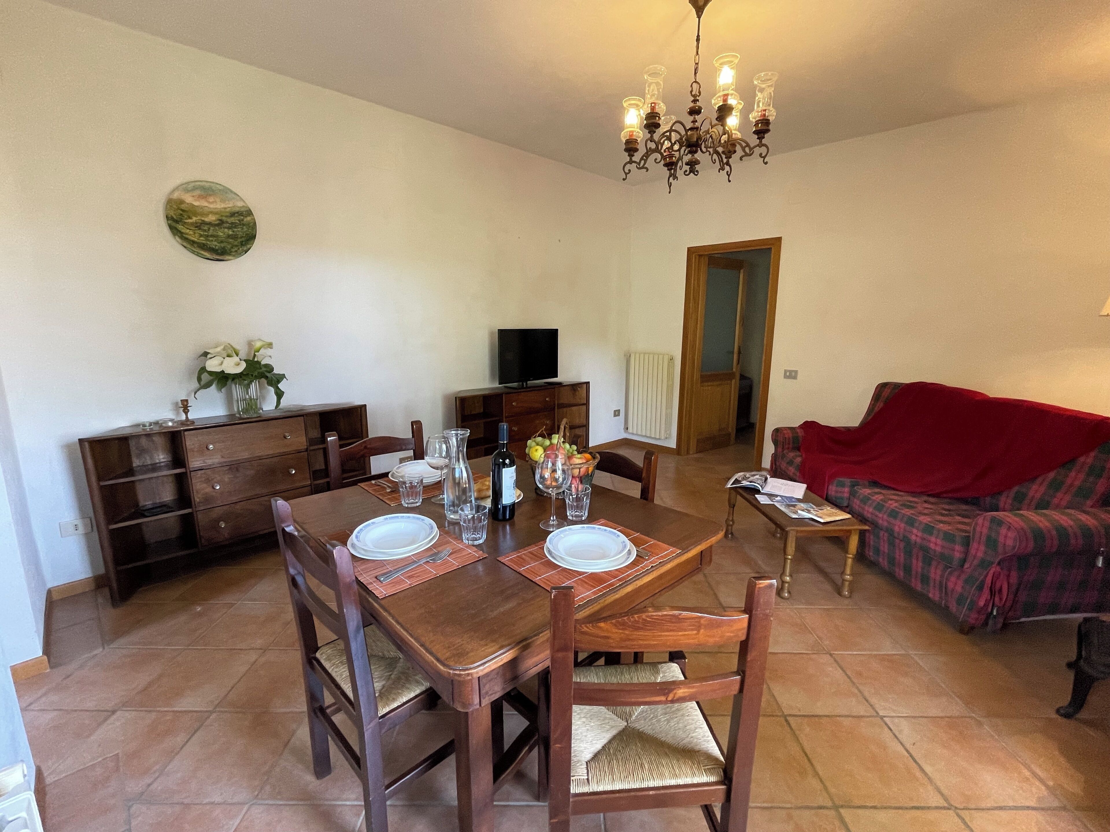 A dining area with a wooden table set for two, a sofa, and a television in the background.