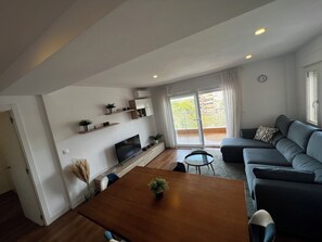 Een moderne woonkamer met een houten eettafel, een bank en een aan de muur bevestigde televisie. Er staan planken met planten en een schuifdeur die leidt naar een balkon.