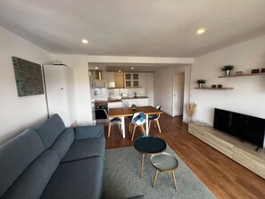 Een moderne woonkamer met een grijze bank, een eettafel met stoelen, een televisie en een keuken op de achtergrond.