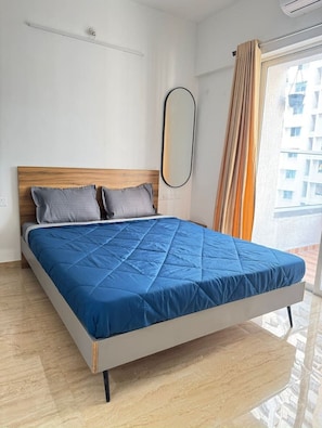 Une chambre à coucher moderne avec une tête de lit en bois, un grand lit recouvert d’une couette bleue, un miroir et une vue sur les bâtiments à travers une fenêtre ornée de rideaux.