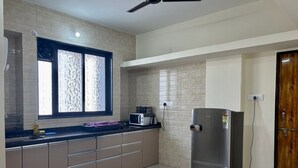 A kitchen with a refrigerator, microwave, and a window with patterned blinds. - Owl's Retreat: 2BHK Modern AC Flat in Wakad, Pune (Pimpri-Chinchwad)