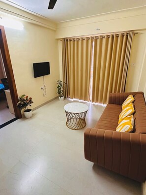A living room with a brown sofa, a round coffee table, a television mounted on the wall, and a large window with curtains.