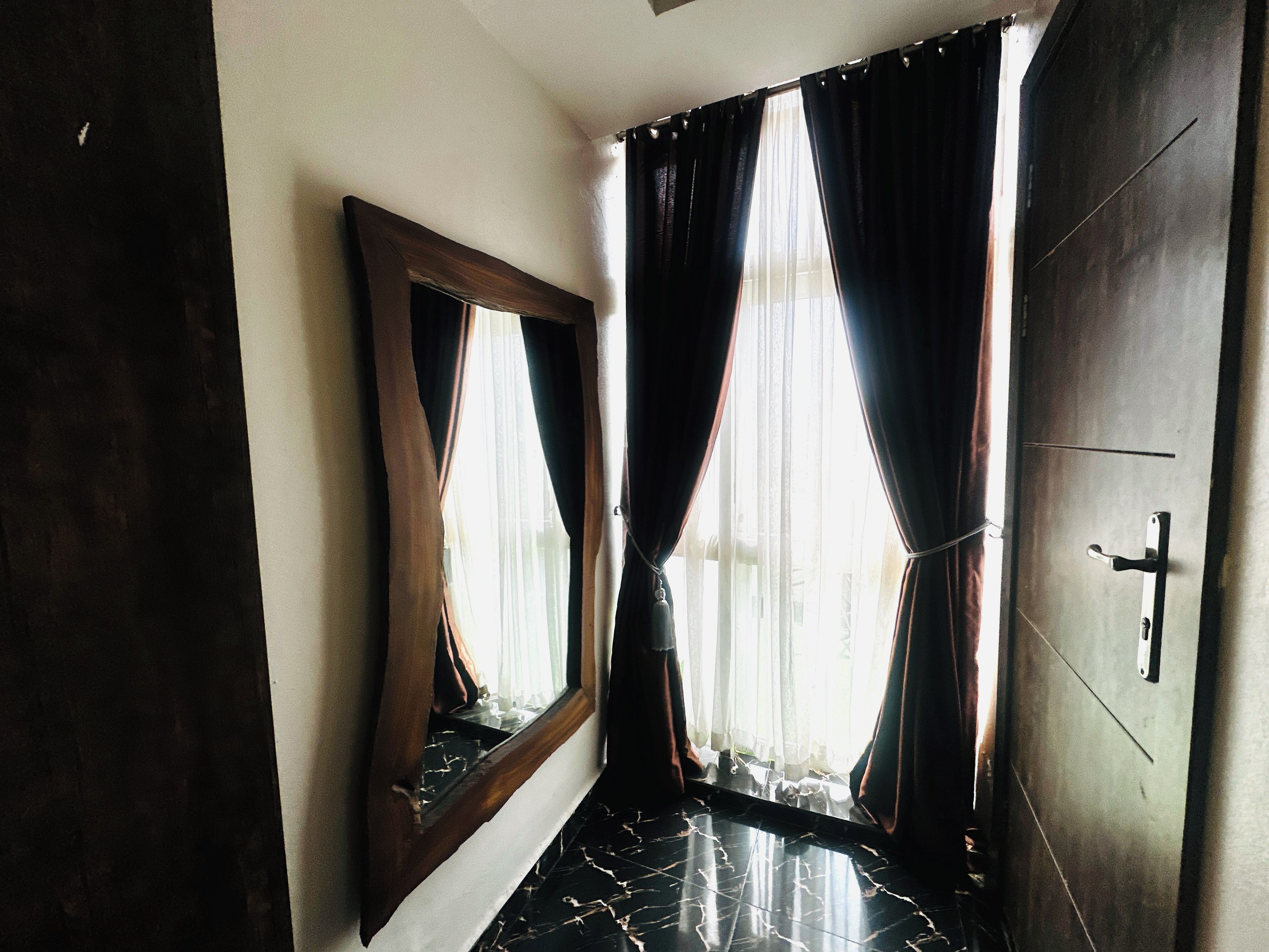 A room with a dark wooden door, a mirror, and a window with sheer curtains.
