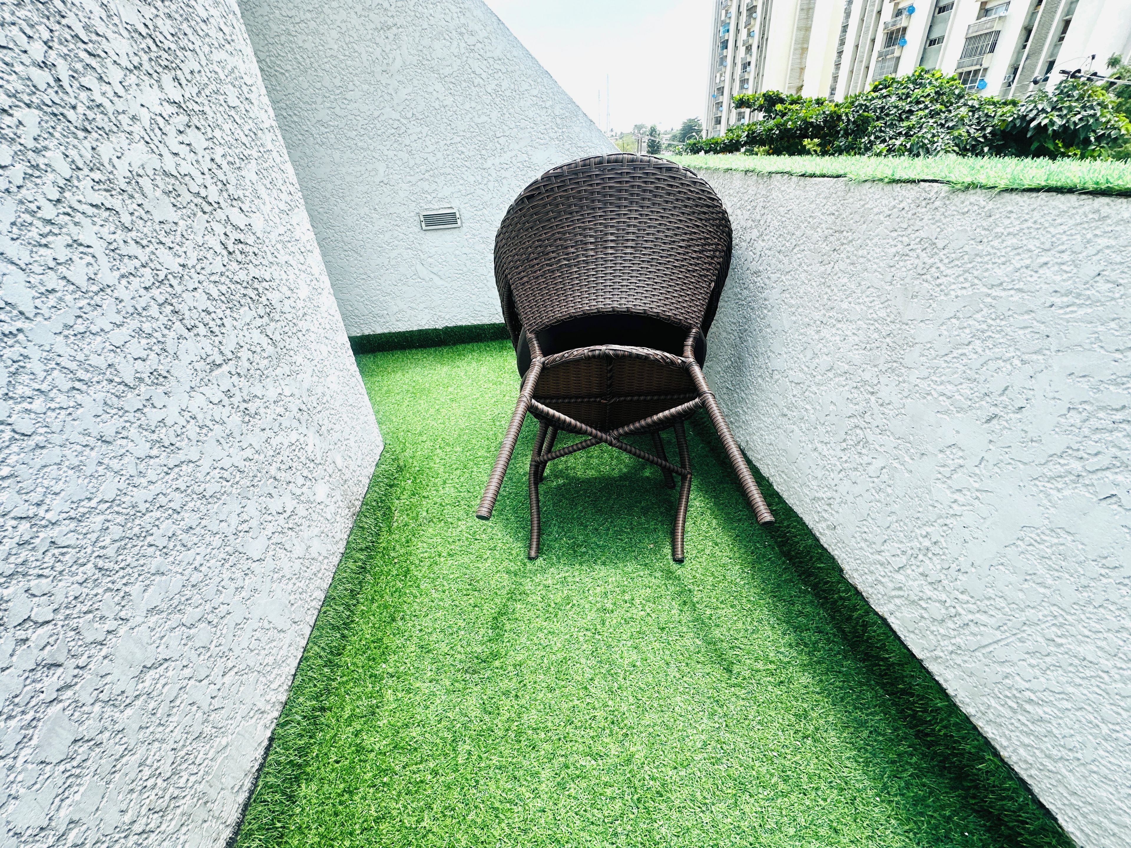 Une chaise en osier posée sur du gazon synthétique, entourée de murs blancs à texture, avec un bâtiment en arrière-plan.