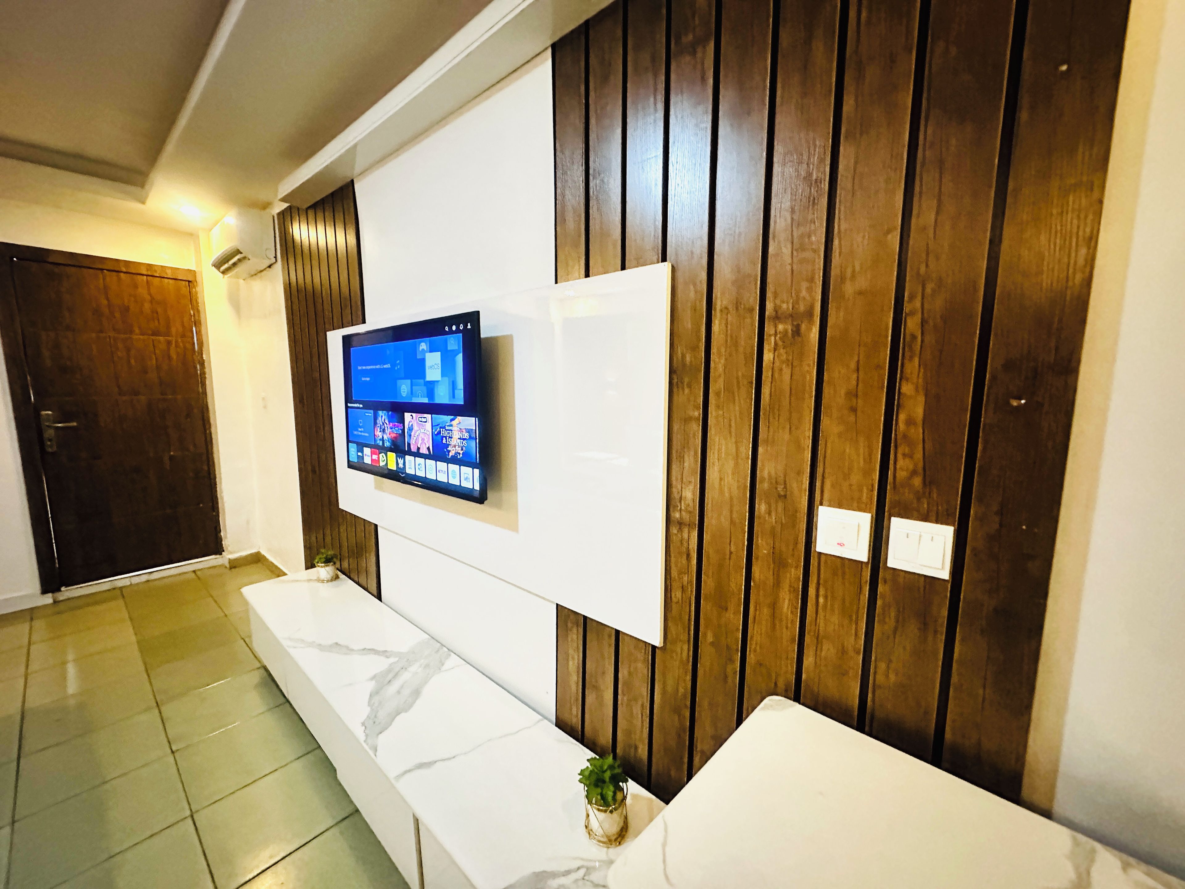 Un lobby moderno de hotel con una televisión de pantalla plana montada en una pared de paneles de madera, un banco de mármol y una puerta.