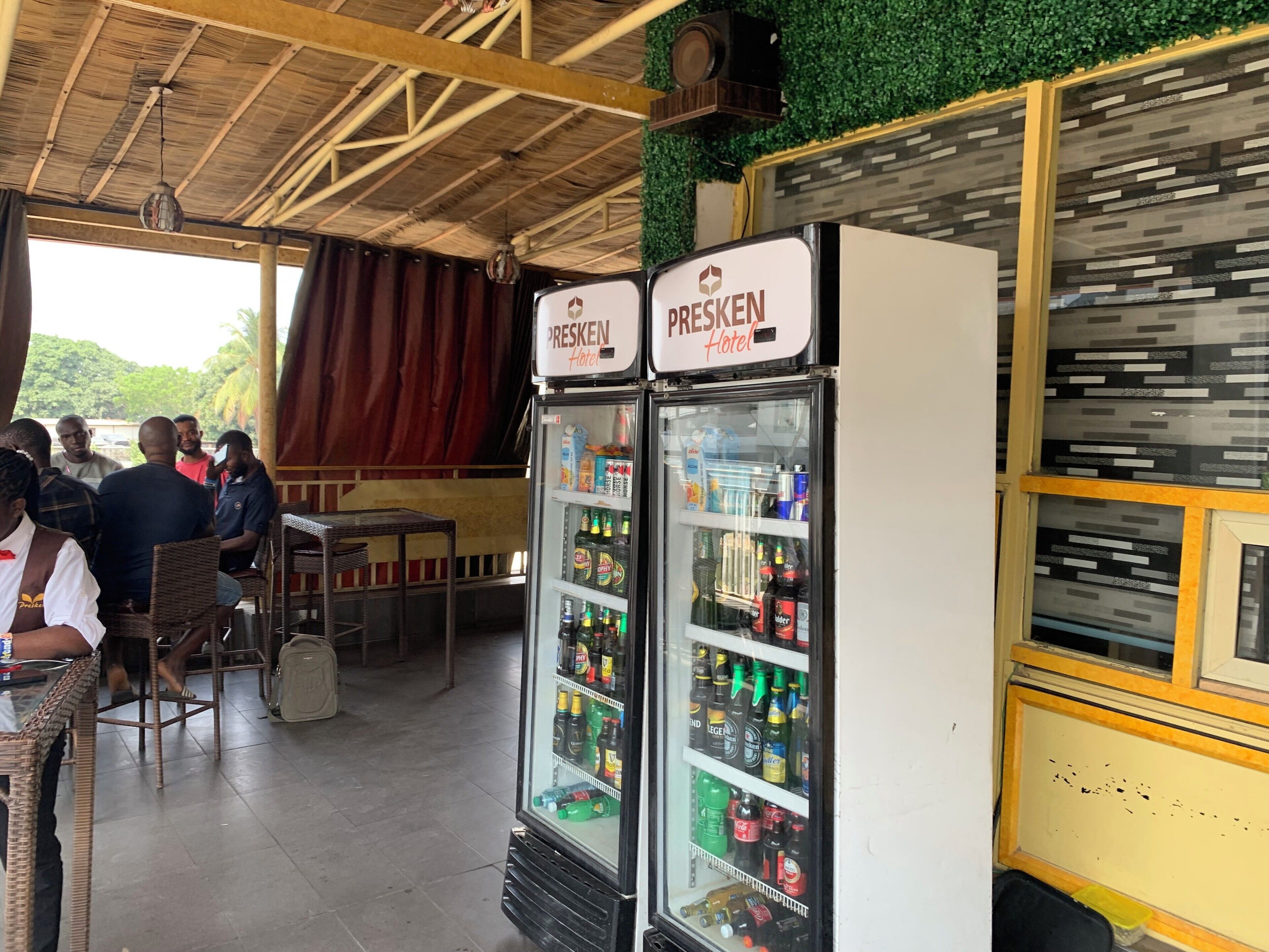 Um saguão de hotel com duas geladeiras identificadas com "PRESKEN Hotel" repletas de bebidas, teto de madeira e parede verde.