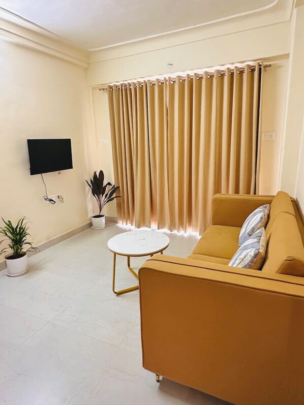 Een woonkamer met een bank, een flatscreen-tv, een klein rond tafeltje en twee potplanten.