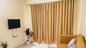 Una sala de estar con un sofá, una mesita redonda, un televisor de pantalla plana montado en la pared y un ventanal con cortinas.