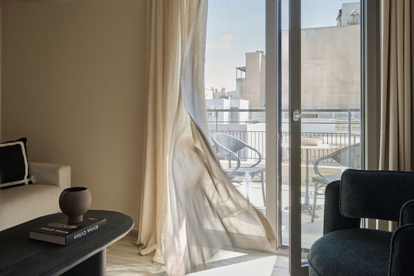 A hotel room with a sofa, a small table with a book and a vase, a chair, and a view of the city through a large window with curtains. - Finders Top Floor Apartments (Athens)