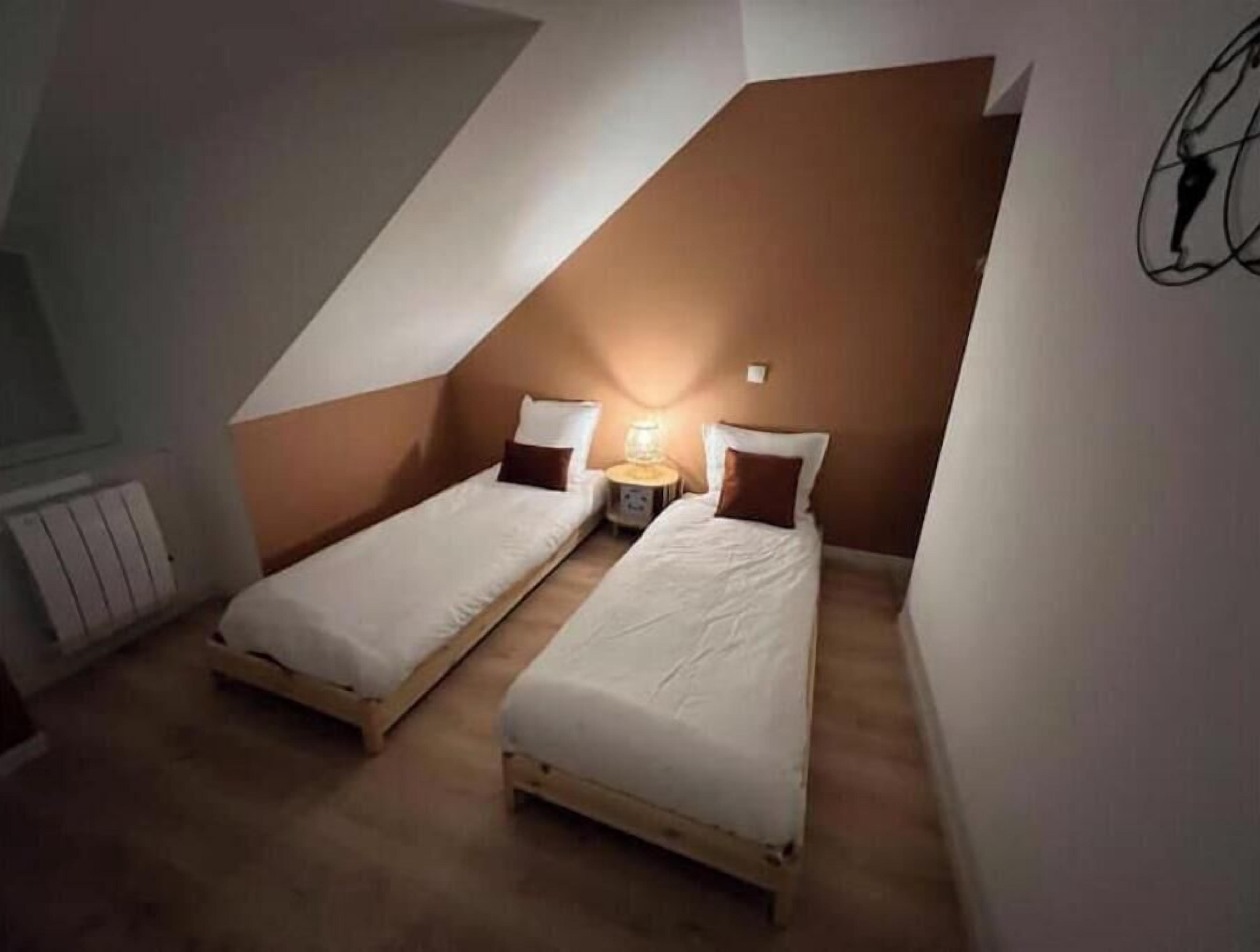 A small, triangular attic bedroom with two single beds, a nightstand, and a wall-mounted lamp.