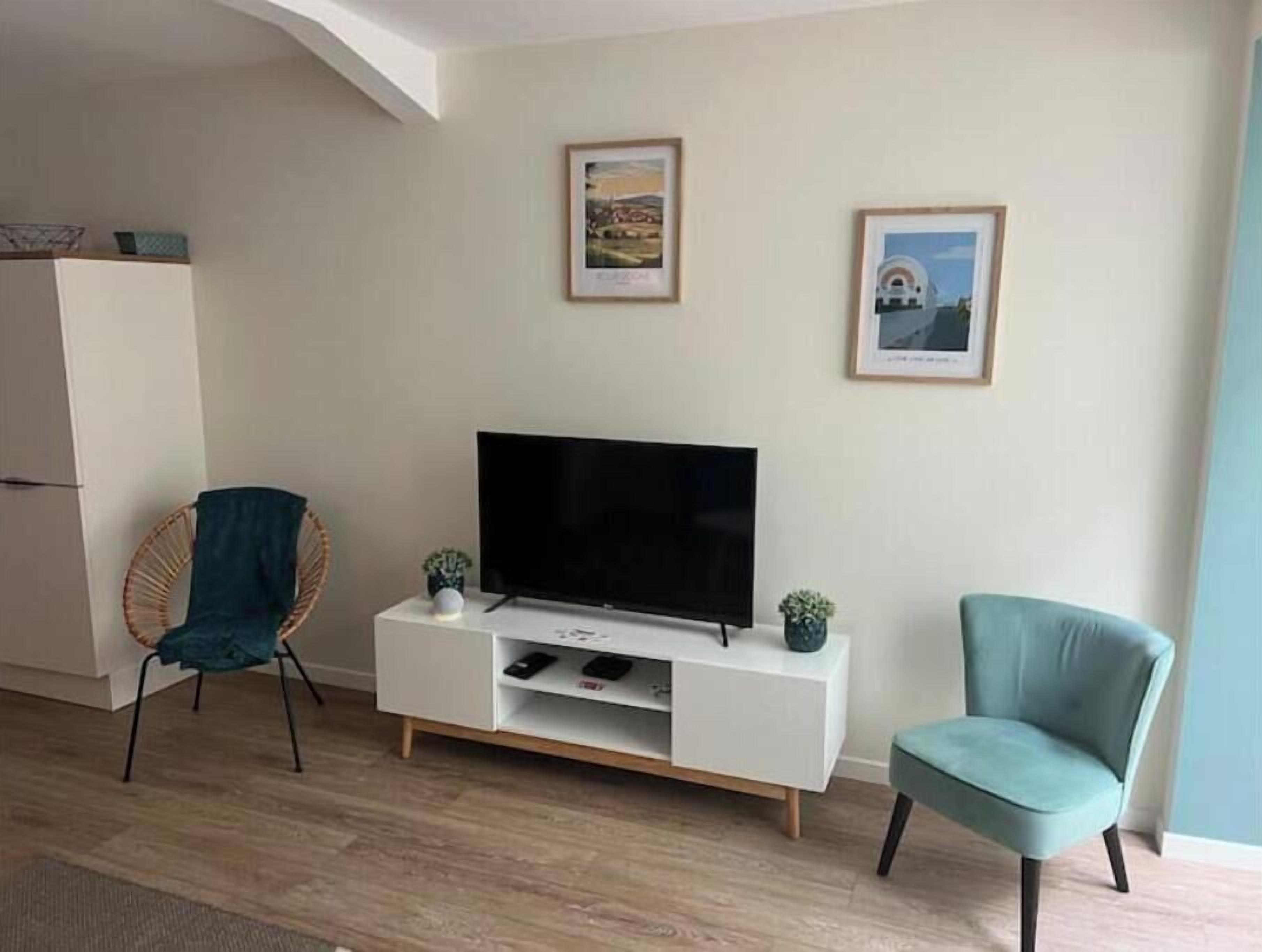 Una sala de estar con un televisor de pantalla plana sobre un mueble blanco, dos cuadros en la pared, una silla de mimbre y un sillón color turquesa.