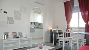 A modern living room with a dining area, white furniture, a small kitchen in the background, and a window with red curtains. - Complete and bright apartment in Monfalcone  (Monfalcone)