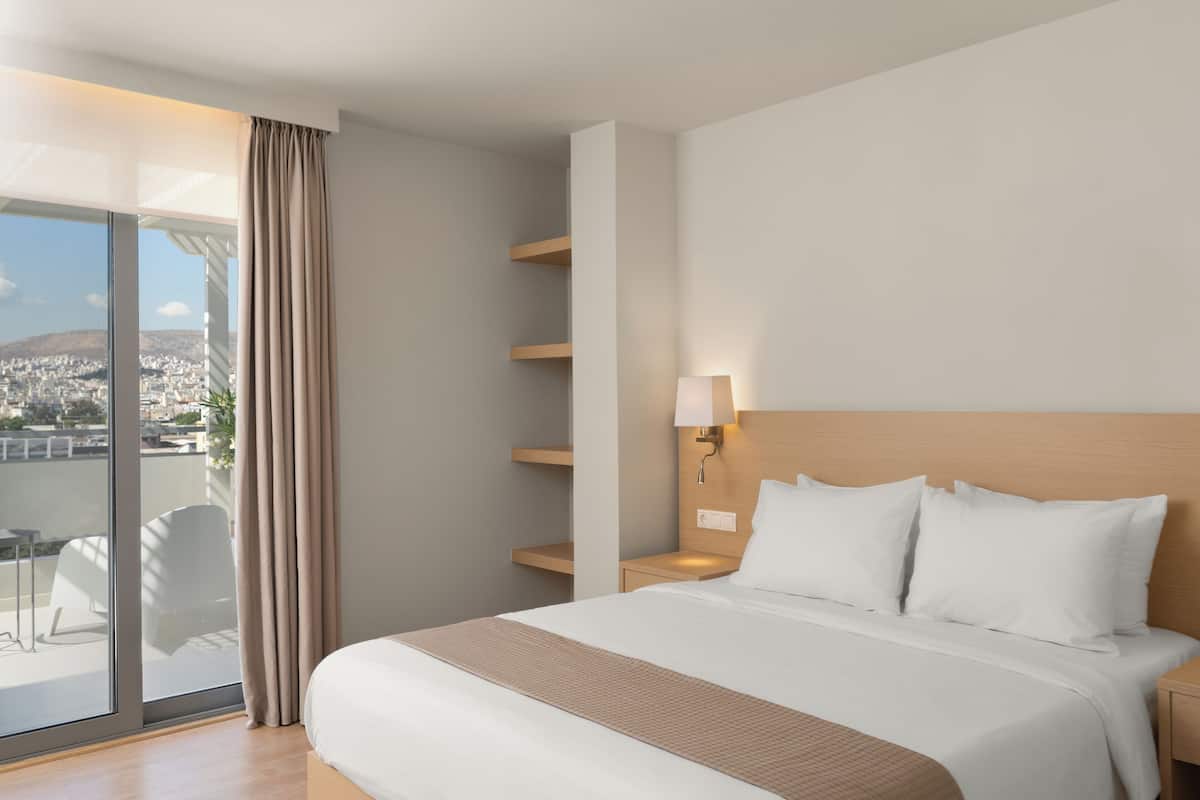 A hotel room with a bed, a nightstand, a lamp, a window with curtains, and a view of the city.