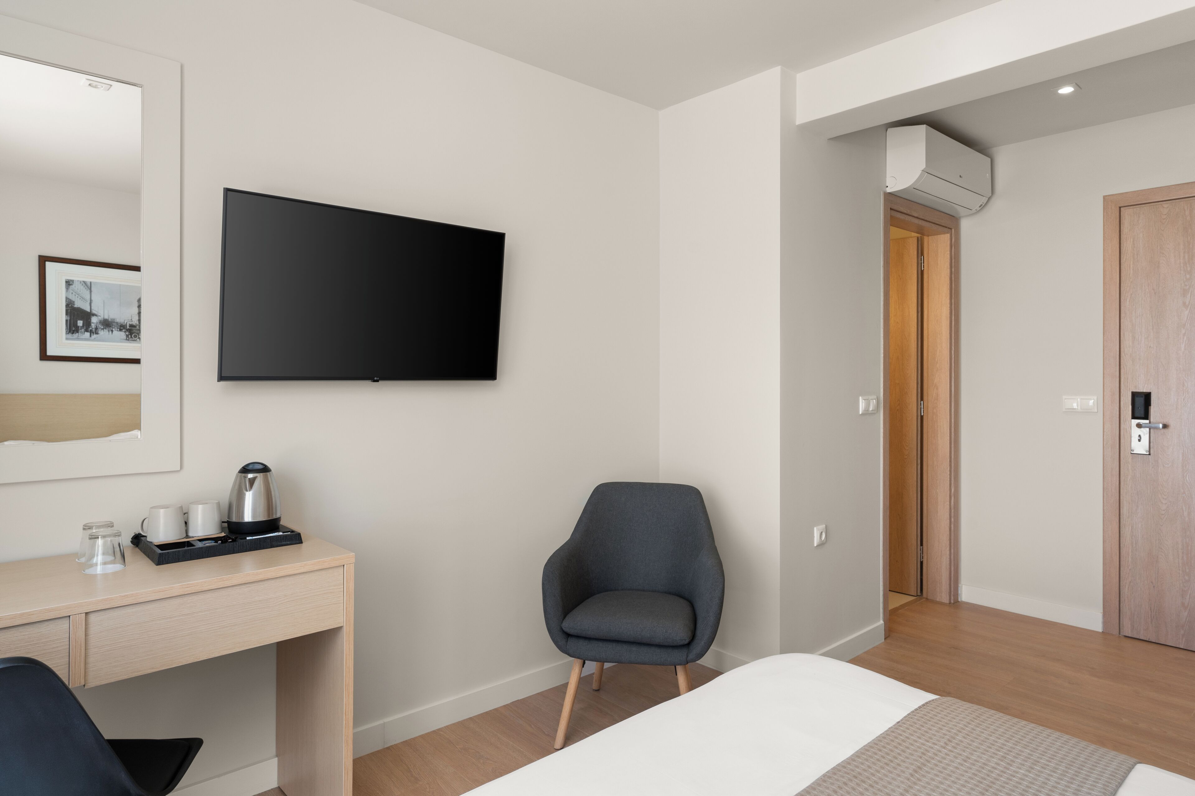 A hotel room with a bed, a desk with a coffee maker, a chair, a TV mounted on the wall, and a door leading to another room.