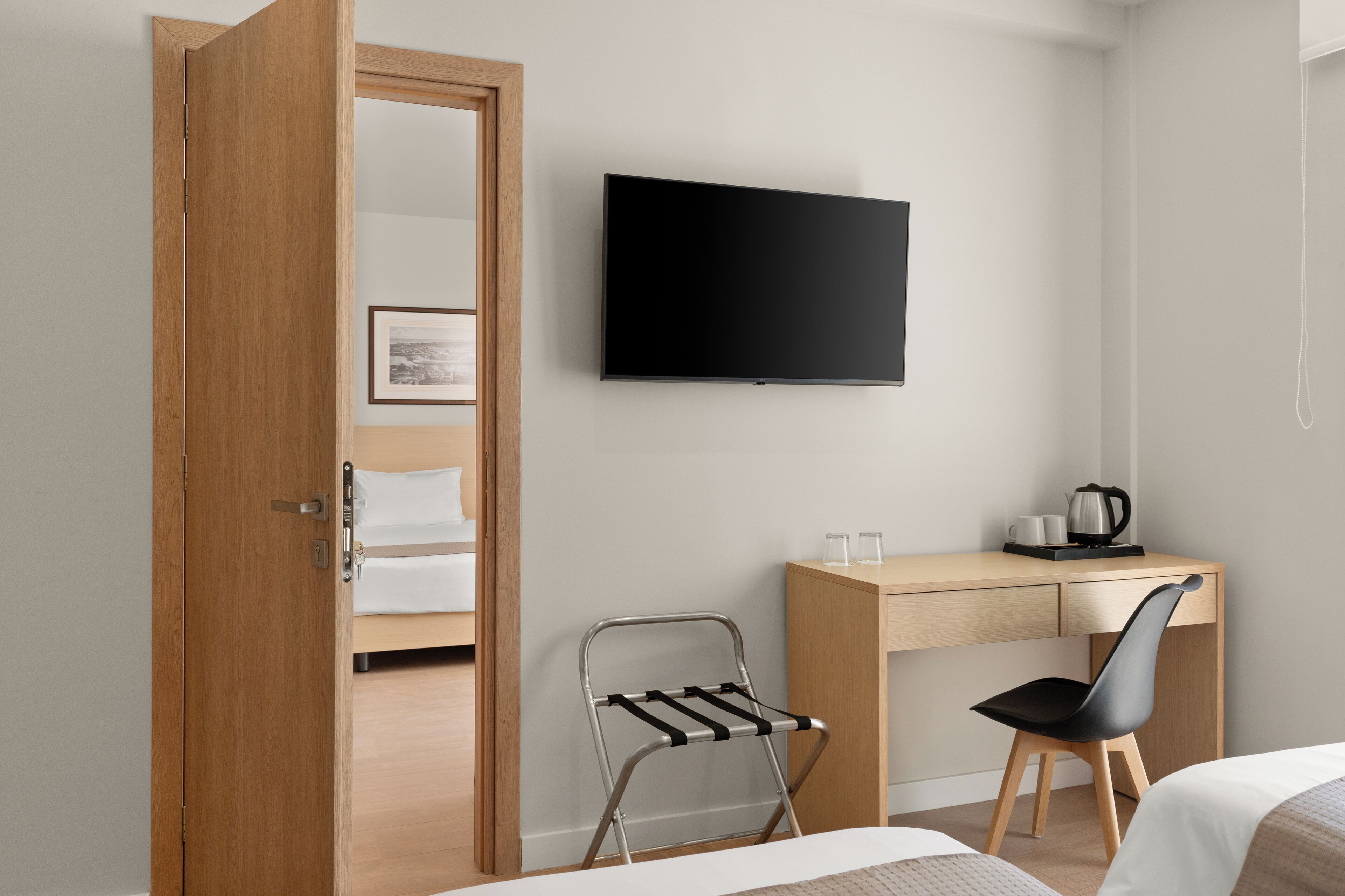 Habitación de hotel con puerta de madera, cama, escritorio con silla, televisor y una mesita con tetera y tazas.