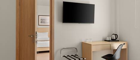 Habitación de hotel con puerta de madera, cama, escritorio con silla, televisor y una mesita con tetera y tazas.