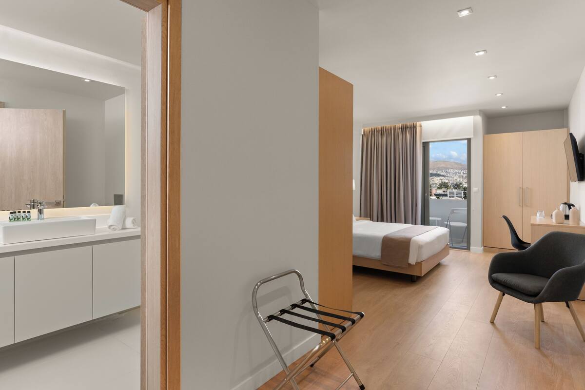 A modern hotel room with a large bed, a sitting area with a chair, a bathroom with a sink and mirror, and a view of the city through the window.