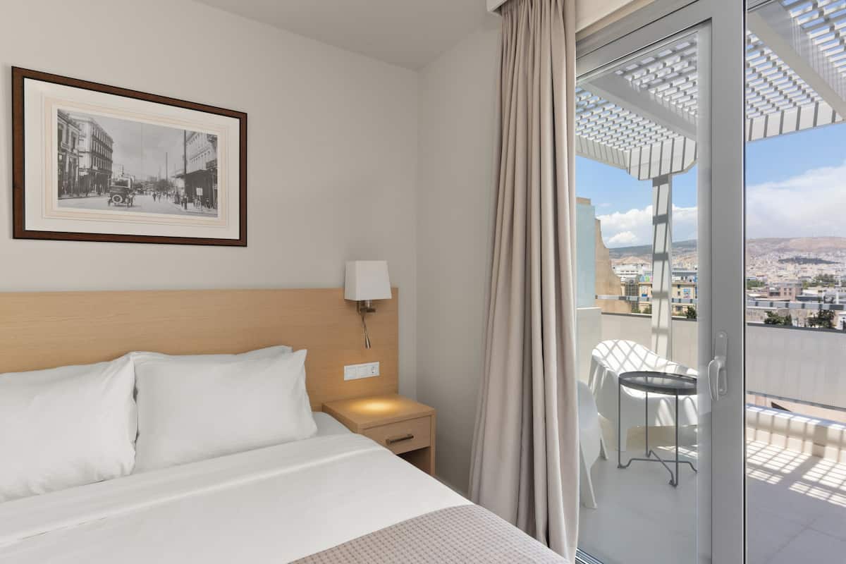A hotel room with a bed, a nightstand, a lamp, a framed picture on the wall, and a balcony with a table and chairs.