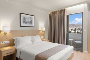 A hotel room with a bed, bedside lamps, a nightstand, a drawer, a framed picture on the wall, and a balcony with a view of the city.