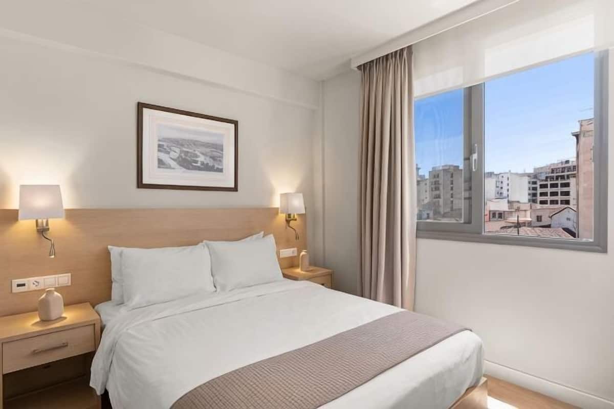 A hotel room with a large bed, two bedside lamps, a framed picture on the wall, and a window with a city view.