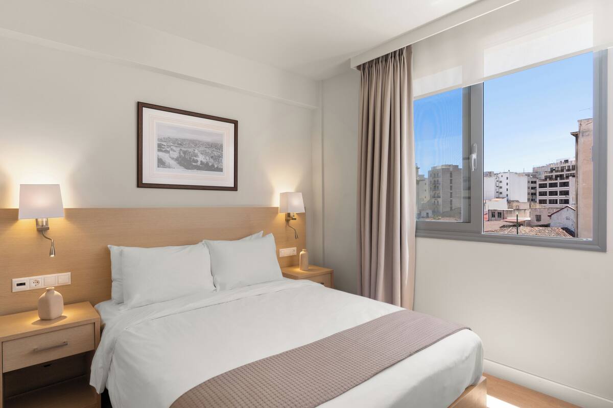 A hotel room with a large bed, two bedside tables, a nightstand, a framed picture on the wall, and a window with a view of buildings.