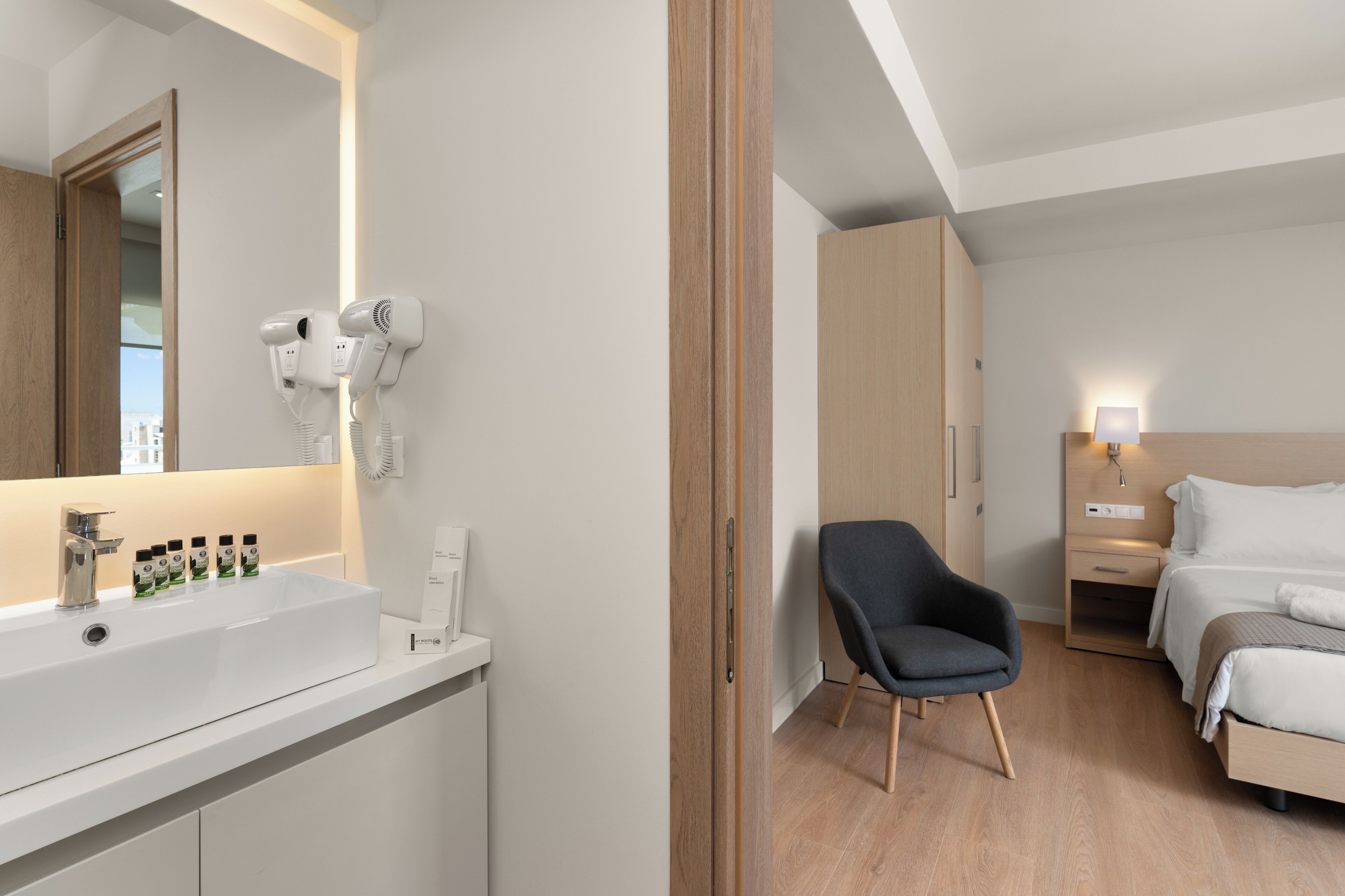 A modern hotel room with a large mirror, a white sink with hairdryers, a bedside table with a lamp, a bed, and a chair.
