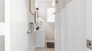 Un pasillo estrecho con una puerta blanca, un refrigerador pequeño de color blanco, un perchero y una ventana con persianas.