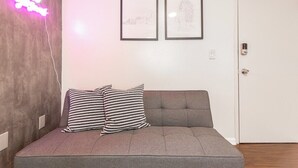 Una habitación con un sofá gris, dos almohadas blancas y negras estampadas, una luz rosa neón y dos obras de arte enmarcadas en la pared.